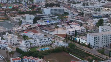 Kıbrıs Rum Kesimi 'nin Ayia Napa kentinin hava görüntüsü, modern oteller, konut binaları, dönme dolap, lunapark ve çevre yeşillikleri gösteriyor.