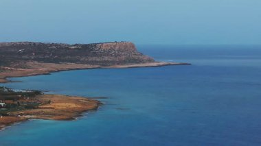 Kıbrıs Rum Kesimi 'nin Ayia Napa kentinin hava görüntüsü, turkuaz suları, kayalık kıyıları, beyazlatılmış otelleri ve kameralar kıyı boyunca hareket ederken dönen tepeleri gösteriyor..