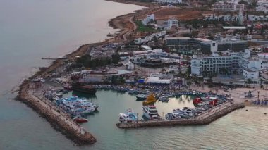 Kıbrıs Rum Kesimi 'nin Ayia Napa havalimanı, mavi sandalyeli kumlu plajlar, tekneleri, kırmızı bayrakları, modern tatil beldeleri ve berrak suları olan bir marina gösteriyor..