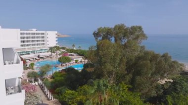 Kıbrıs Rum Kesimi 'nin Ayia Napa kentindeki bir kıyı bölgesinin havadan görünüşü, bir bahçe, mavi şemsiyeli kumlu plaj, palmiye ağaçları ve Akdeniz' i gösteriyor..