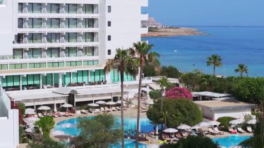 Kıbrıs Rum Kesimi 'nin Ayia Napa kentinin hava manzarası, berrak mavi sular, palmiye ağaçları, şezlonglu bir havuz ve sahil şeridi manzarasını gösteriyor..