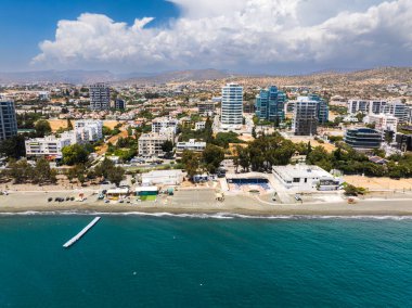 Kıbrıs Rum Kesimi 'nin Limasol kentinin havadan görünüşü, yüksek binalar, kumlu plaj, turkuaz sular, küçük bir iskele, yeşil alanlar ve kayan tepeleri gösteriyor..