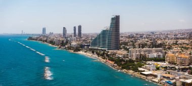Kıbrıs 'ın Limasol kentinin havadan görünüşü, modern yüksek binalar, eğimli bir yapı, turkuaz deniz, kumlu plaj ve dalga kırıcılara sahip..