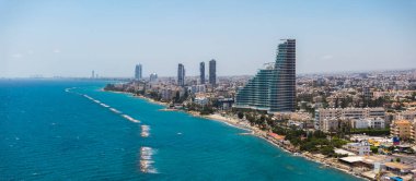 Kıbrıs Rum Kesimi 'nin Limasol kentinin havadan görünüşü, kıyı yakınlarındaki modern yüksek binalar, berrak mavi sular, yerleşim alanları ve vinçli uzak bir liman.