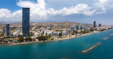 Kıbrıs 'ın Limasol kentinin havadan görünüşü, modern gökdelenleri, karanlık cam bir kuleyi, Akdeniz kıyı şeridini, dalgaları, tepeleri ve parçalı bulutlu gökyüzünü gösteriyor..