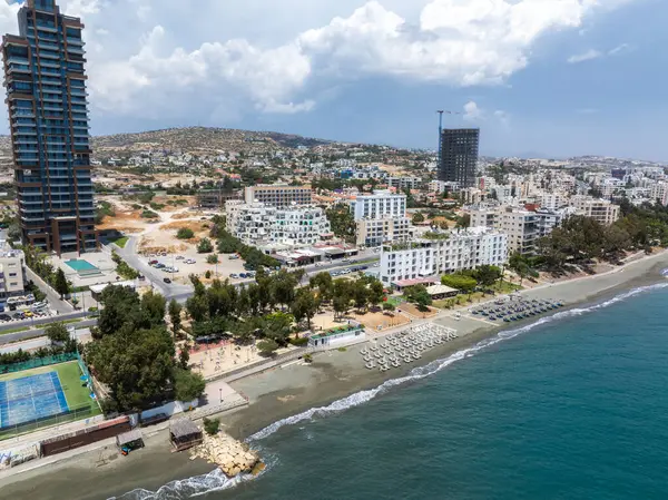 Kıbrıs 'ın Limasol kentinin havadan görünüşü, kumlu bir sahil, güneşli şezlonglar, berrak Akdeniz suları, modern yüksek binalar, yeşillikler ve uzak tepeler yer alıyor..