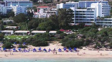 Kıbrıs Rum Kesimi 'nin Ayia Napa kentinin hava manzarasında beyaz binalar, yemyeşil yemyeşil alanlar, güneşli bir kumsal ve turkuaz Akdeniz suları görülüyor..