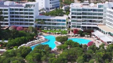 Kıbrıs Rum Kesimi 'nin Ayia Napa kentindeki bir tatil beldesinde geniş bir havuz, güneş pansiyonları, modern beyaz binalar ve palmiye ağaçlarıyla canlı manzaralar görülüyor..