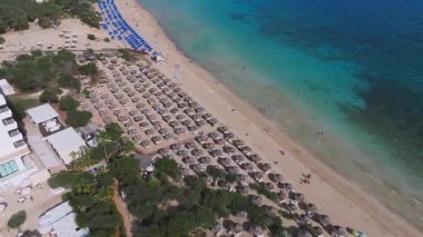 Kıbrıs Rum Kesimi 'nin Ayia Napa kentindeki bir plajın havadan görünüşü, sıralanmış güneş yatakları, mavi şemsiyeler, turkuaz sularda yüzenler ve kıyı şeridinde bir tatil köyü gösteriyor..