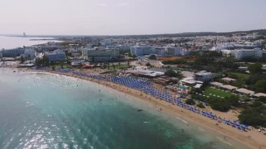 Ayia Napa 'nın güneşli yatakları, şemsiyeleri ve berrak sularıyla kumlu plajının havadan görüntüsü. Havuzu ve yeşilliği olan oteller görülebiliyor, tepeleri uzakta..