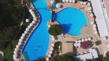 Kıbrıs Rum Kesimi 'nin Ayia Napa kentindeki bir tatil köyünün hava manzarasında, ahşap köprüsü, beyaz şemsiyeleri, şezlongları ve yemyeşil yeşillikleri olan eşsiz bir havuz yer alıyor..
