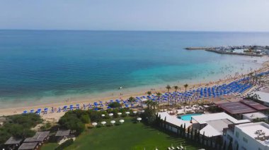 Ayia Napa 'nın Kıbrıs' taki kıyı şeridini gösteren hava manzarasında, mavi şemsiyeli kumlu bir sahil, havuzlu bir tatil beldesi ve hareket halinde bir sürat teknesi yer alıyor..