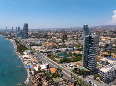 Kıbrıs 'ın Limasol kentinin havadan görünüşü, modern yüksek binaları, eğimli binaları, kıyı yeşilliklerini, ana yolları ve mavi gökyüzünün altındaki uzak tepeleri gösteriyor..