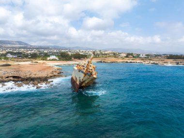Edro III Batığı, Kıbrıs 'ın turkuvaz sularıyla çevrili kayalık kıyıları yakınında uzanırken, arka planda bir kasaba ve dalgalı tepeler görülebiliyor..