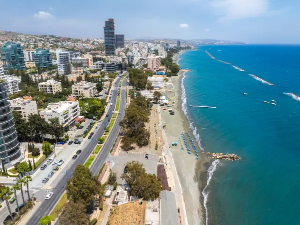 Kıbrıs 'ın Limasol kentinin havadan görünüşü, modern binalar, kıyı yolu, şemsiyeli kumlu plaj, turkuaz deniz ve güneşli gökyüzü altında küçük rıhtımlar gösteriyor..