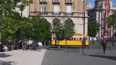 Budapeşte 'de bir binanın yanında tarihi bir tramvay, süslü mimarisi olan bir plaza, ağaçlar, banklar, yürüyen ve etkileşen insanlar, ve arka planda kırmızı bir vinç..