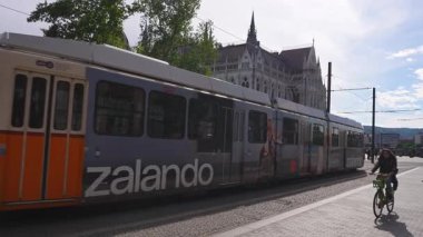 Budapeşte 'deki Macar Parlamentosu' nun yeşil ağaçlar, taş bir heykel ve kaldırım taşı bir sokak tarafından çerçevelenmiş turuncu bir tramvayla görüntüsü.