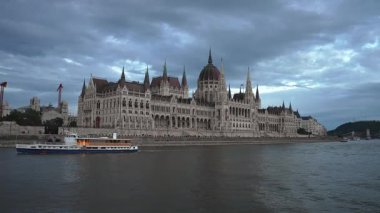 Macaristan 'ın Budapeşte kentindeki Tuna Nehri' nde bir tekne süzülüyor ve arkasında Macar Parlamento Binası ve bulutlu bir gökyüzü bulunuyor..