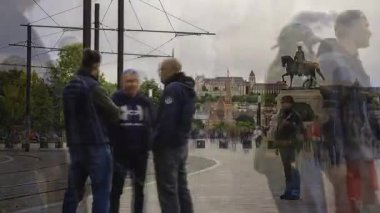 Zaman çizelgesi, arka planda Buda Kalesi olan bir binicilik heykelinin yanında hareket eden yayaları gösteriyor. Tramvay hatları ve kentsel öğeler görünür.