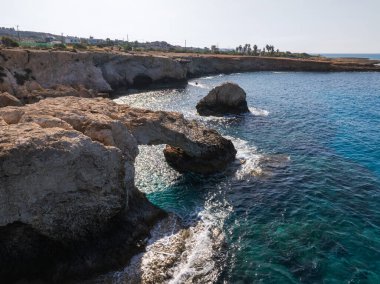 Kıbrıs 'ın Ayia Napa kentindeki kıyı sahnesinde, berrak mavi suların üzerinde bir kaya kemeri, engebeli kıyı şeridi, küçük mağaralar ve arka planda seyrek bitki örtüsü bulunuyor..