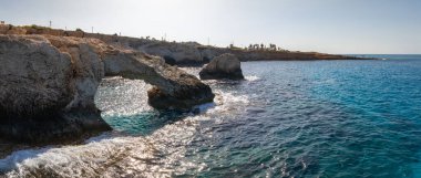 Kıbrıs 'ın Ayia Napa kentindeki kıyı manzarası, mavi suların üzerinde bir kaya aşk kemeri, engebeli uçurumlar, ufukta dağılmış ağaçlar ve yapılar.