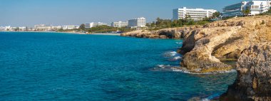 Engebeli bej kayalıklar Kıbrıs Rum Kesimi 'nin Ayia Napa kentindeki turkuaz Akdeniz sularıyla, kumlu plajları, güneşli salları ve yakınlarındaki modern lüks otellerle karşılaşıyor..