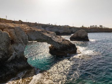 Doğal bir kaya kemeri Ayia Napa kıyı şeridi boyunca uzanan turkuaz suları, engebeli uçurumlar, dağınık ağaçlar ve güneş ışığıyla dalgaları yansıtır..