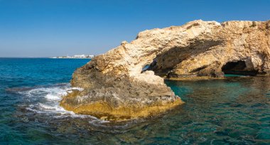 Kıbrıs 'ın Ayia Napa kenti yakınlarında turkuaz sularla çevrili doğal bir kireçtaşı Aşk kemeri. Parlak mavi gökyüzü ve görünen uzak beyaz binaları var..