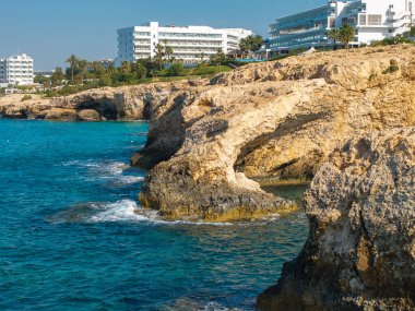 Kireçtaşı kayalıkları Kıbrıs 'ın Ayia Napa kentindeki turkuaz Akdeniz sularıyla buluşurken, arka planda çağdaş beyaz tatil beldeleri, palmiye ağaçları ve yeşillikler yer alıyor..