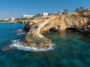 Turkuaz sular Kıbrıs Rum Kesimi 'nin Ayia Napa kentinde doğal kemerli, engebeli kireçtaşı kayalıklarla buluşurken, arka planda lüks oteller ve palmiye ağaçları bulunuyor..