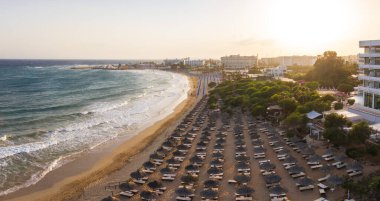 Günbatımında Kıbrıs 'taki Ayia Napa plajının hava manzarasında turkuaz sular, güneş pansiyonları, saman şemsiyeler, tatil beldeleri ve uzak bir kasaba manzarası yer alıyor..