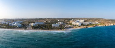 Kıbrıs 'ın Ayia Napa kentinin hava manzarasında turkuaz sular, kumlu plajlar, lüks oteller, lüks oteller, yemyeşil alanlar ve açık mavi bir gökyüzü görülüyor..