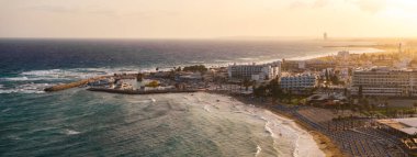 Kıbrıs Rum Kesimi 'nin Ayia Napa kentinin gün batımında kumlu sahilleri, turkuaz suları, arka planda tekneleri ve lüks otelleri olan küçük bir liman manzarası..