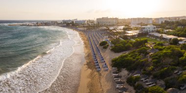 Kıbrıs Rum Kesimi 'nin Ayia Napa kentindeki kumlu sahil manzarasında mavi güneş pansiyonları, turkuaz dalgalar, yemyeşil, sazdan kabinler ve lüks oteller yer alıyor..