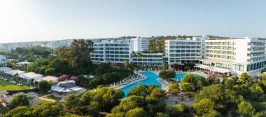 Modern lüks otel, yemyeşil, canlı çiçekler, ve dinlenme yerlerinin olduğu büyük bir havuz. Arka planda kentsel ve doğal manzaralar.