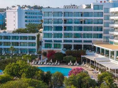 Camdan pencereleri ve balkonları olan modern çok katlı bir otel, güneşli havuzlu merkezi bir havuz, canlı bir yeşillik ve Ayia Napa 'da açık mavi bir gökyüzü..