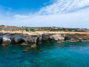 Doğal deniz mağaraları olan engebeli kireçtaşı kayalıkları, arka planda dağınık binalarla Kıbrıs 'ın Ayia Napa kentindeki turkuaz Akdeniz sularıyla buluşuyor.