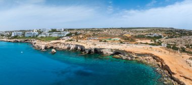 Turkuaz Akdeniz suları Kıbrıs Rum Kesimi 'nin Ayia Napa kentinde kireçtaşı kayalıklarıyla buluşuyor. Arka planda lüks oteller, asfaltlı bir yol ve seyrek bitki örtüsü görülebilir..