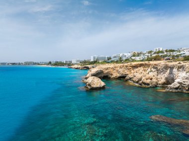 Kıbrıs Rum Kesimi 'nin Ayia Napa kentindeki turkuaz sular ve kayalık uçurumlar, modern beyaz lüks oteller ve arka planda açık mavi bir gökyüzünün altında yeşillik.