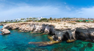Deniz mağaralı engebeli kireçtaşı kayalıkları Kıbrıs Rum Kesimi 'nin Ayia Napa kentinde turkuaz sularla buluşuyor. Arka planda beyaz lüks tatil beldeleri ve yeşillikler görülüyor.