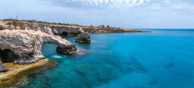 Kıbrıs Rum Kesimi 'nin Ayia Napa kentindeki turkuaz Akdeniz sularına uzanan kireçtaşı kayalıkları ve doğal kaya kemerleri açık bulutlu açık bir gökyüzü altında.