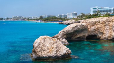 Kıbrıs 'taki Ayia Napa kıyı şeridi berrak turkuaz sular, engebeli kayalar, kumlu plajlar, güneşli banyolar ve arka planda modern lüks oteller.