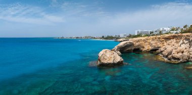 Kıbrıs Rum Kesimi 'nin Ayia Napa kentindeki turkuaz sular ve engebeli kaya oluşumları, kıyı şeridi boyunca berrak bir gökyüzü altında modern beyaz oteller ve tatil beldeleri bulunuyor..
