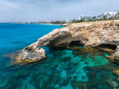 Kıbrıs Rum Kesimi 'nin Ayia Napa kentindeki turkuaz suların üzerinde doğal kaya aşk kemeri, görünür deniz yatakları ve arka planda kıyı şeridini kaplayan modern beyaz oteller bulunuyor..