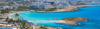 Kıbrıs Rum Kesimi 'nin Ayia Napa kentindeki Nissi Sahili' nin havadan görünüşü, turkuaz sular, güneş pansiyonları, yüzücüler, yakınlardaki tatil beldeleri, yemyeşil alanlar ve kayalık bir adacık..