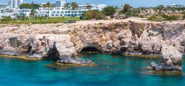 Doğal mağaraları olan engebeli kireçtaşı kayalıkları, arka planda bir yüzücü ve lüks tatil beldeleri bulunan Kıbrıs Rum Kesimi 'nin Ayia Napa kentindeki berrak turkuaz sularıyla buluşuyor..