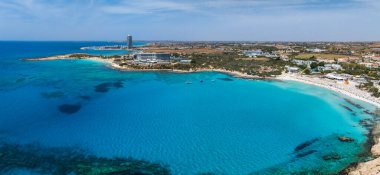 Kıbrıs 'ın Ayia Napa kentindeki Nissi Sahili' nin havadan görünüşü, turkuaz sular, beyaz kum, lüks tatil beldeleri, yüksek bir kule, yemyeşil ve küçük tekneler..