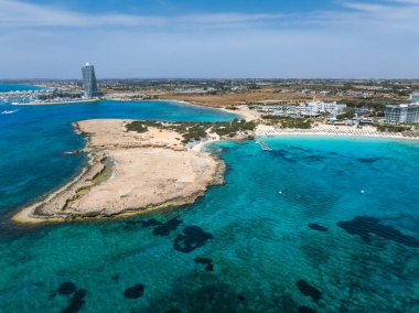 Kıbrıs Rum Kesimi 'nin Ayia Napa kentindeki Nissi Plajı' nın turkuaz sular, kumlu kıyılar, güneşli yataklar, şemsiyeler, lüks tatil beldeleri ve modern bir gökdelen binası ile hava manzarası.