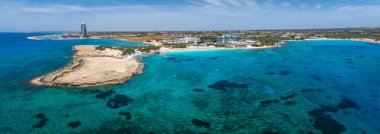 Kıbrıs 'ın Ayia Napa kentinin havadan görünüşü, Nissi ve Landa sahillerini, turkuaz suları, kayalık bir yarımada, lüks tatil beldeleri ve modern bir kıyı kulesini gösteriyor..