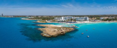 Kıbrıs Rum Kesimi 'nin Ayia Napa kentindeki Nissi Plajı' nın turkuaz sular, kumlu kıyılar, lüks tatil beldeleri, güneş pansiyonları ve yüzen insanlarla hava manzarası.